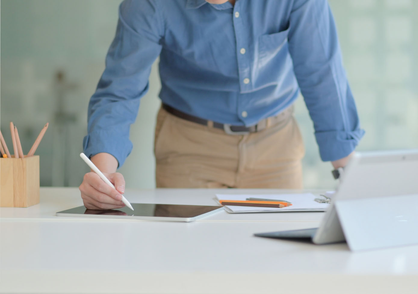 A man stands in front of desk, working with a digital pen on a tablet while reviewing work on a fold-out laptop.