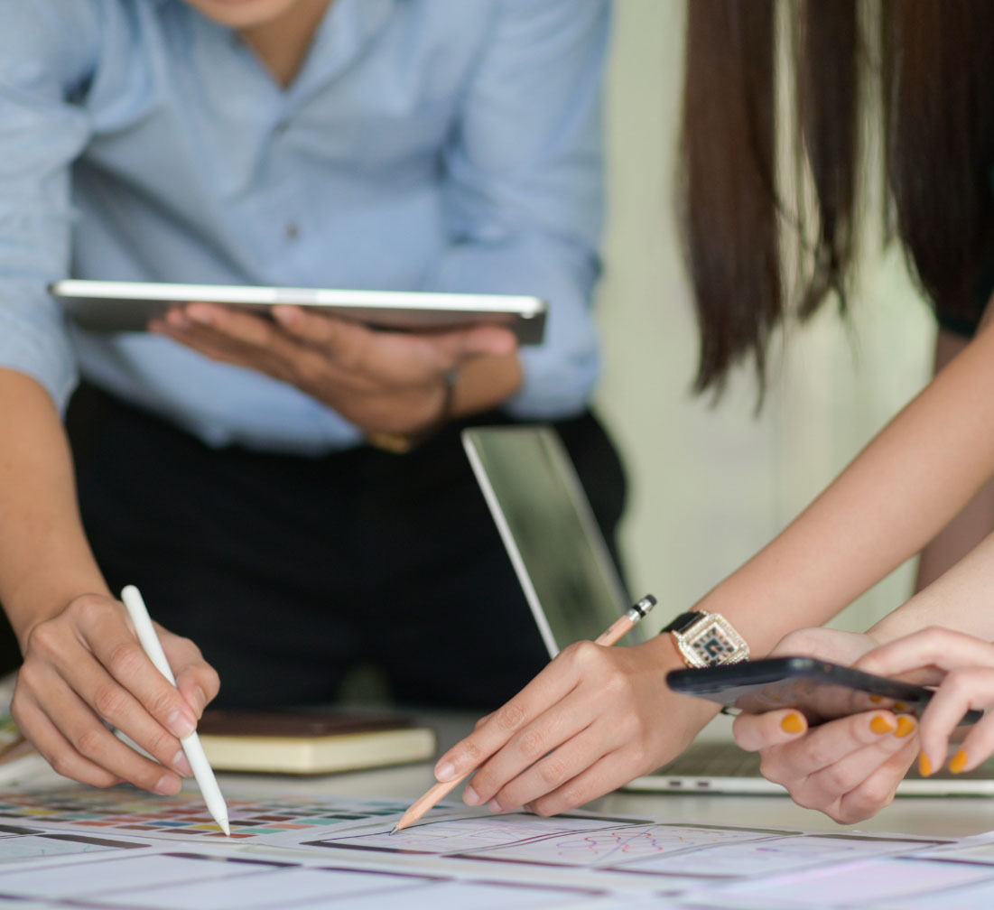 Two people each holding an electronic device and an pencil pointing a printout on the table