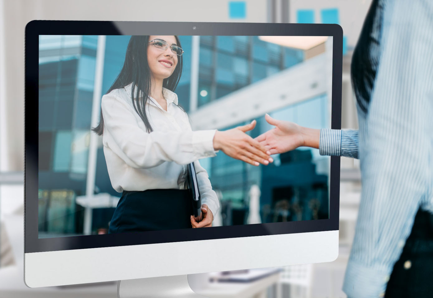 A professional woman reaches out her hand from inside a monitor to shake hands with another professionally dressed person.