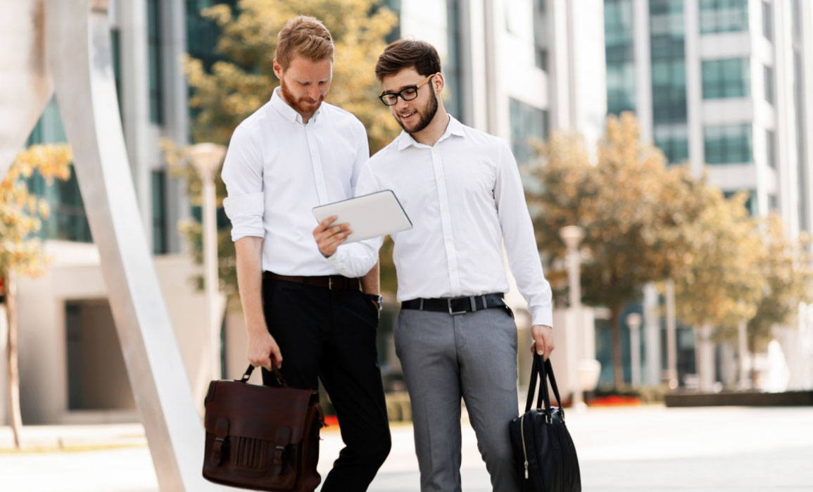 Two IT professionals walking out of an office building park, looking to be an tablet with smiles on their faces.