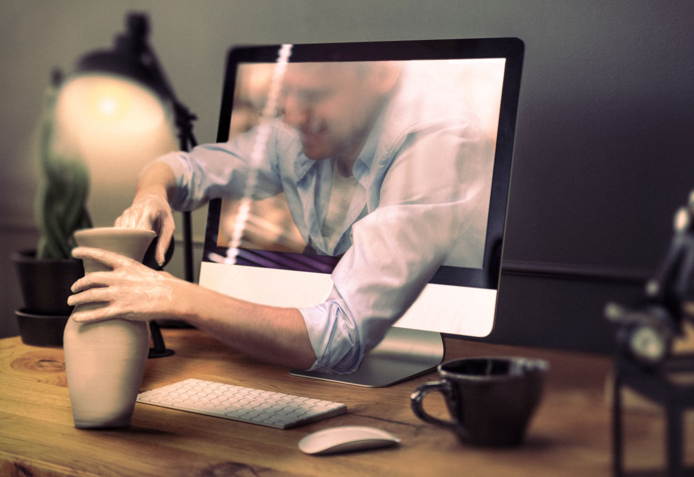 A digital man extends his arms out of a computer monitor's screen, which is sitting on a desk with a keyboard, mouse and mug, and is handcrafting a clay jar.