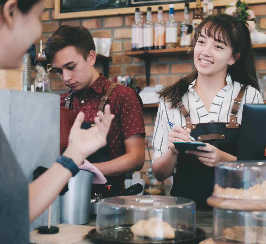 A young lady smiles while working behind the counter at a shop, listening and taking an order while conversing with a customer.