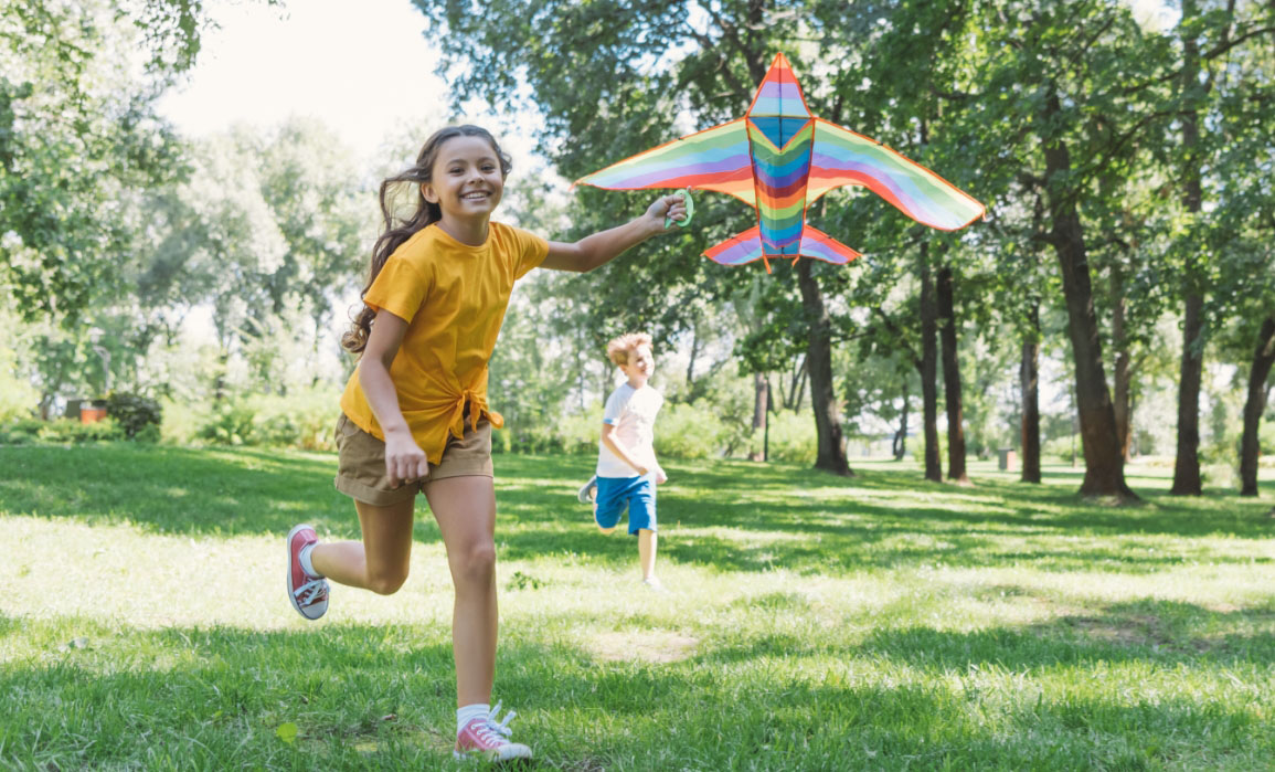 A young girl runs through a park while flying a rainbow-colored kite shaped like a bird and being chased by her younger brother.