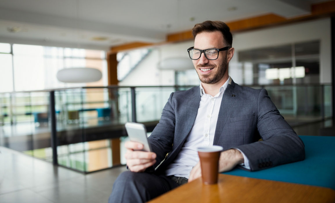 A man, dressed business casual, sits at a table in front of his coffee, looking at his phone and smiling and what he is seeing.