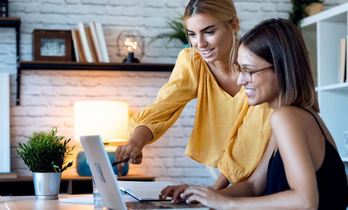 A woman is smiling and seated in from of a laptop at a desk, while another woman, who is smiling and standing next to her, points to the screen of the laptop with a pen.