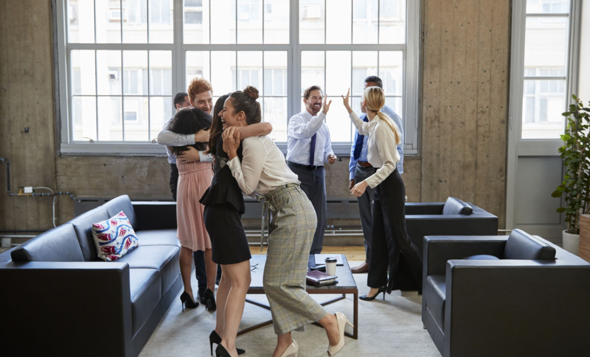 A group of professional men and women have stood up from their informal meeting, which took place on couches in a lobby, and are celebrating together. Some of the people are hugging, others high-five each other.